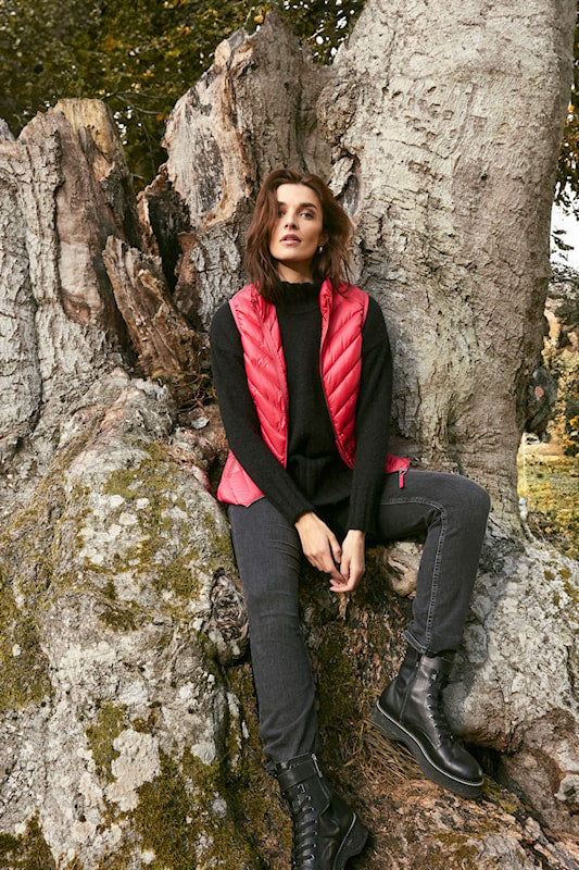 A woman sits on a large tree stump outdoors, wearing a bright pink quilted puffer vest over a black turtleneck sweater, dark gray jeans, and black lace-up boots.