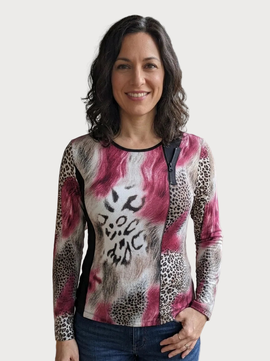 A woman with dark shoulder-length hair stands indoors wearing a long-sleeve zip-front top with a pink, black, and leopard-print abstract pattern, paired with blue jeans. The background shows a softly blurred living space with a brick column and a light-colored sofa.