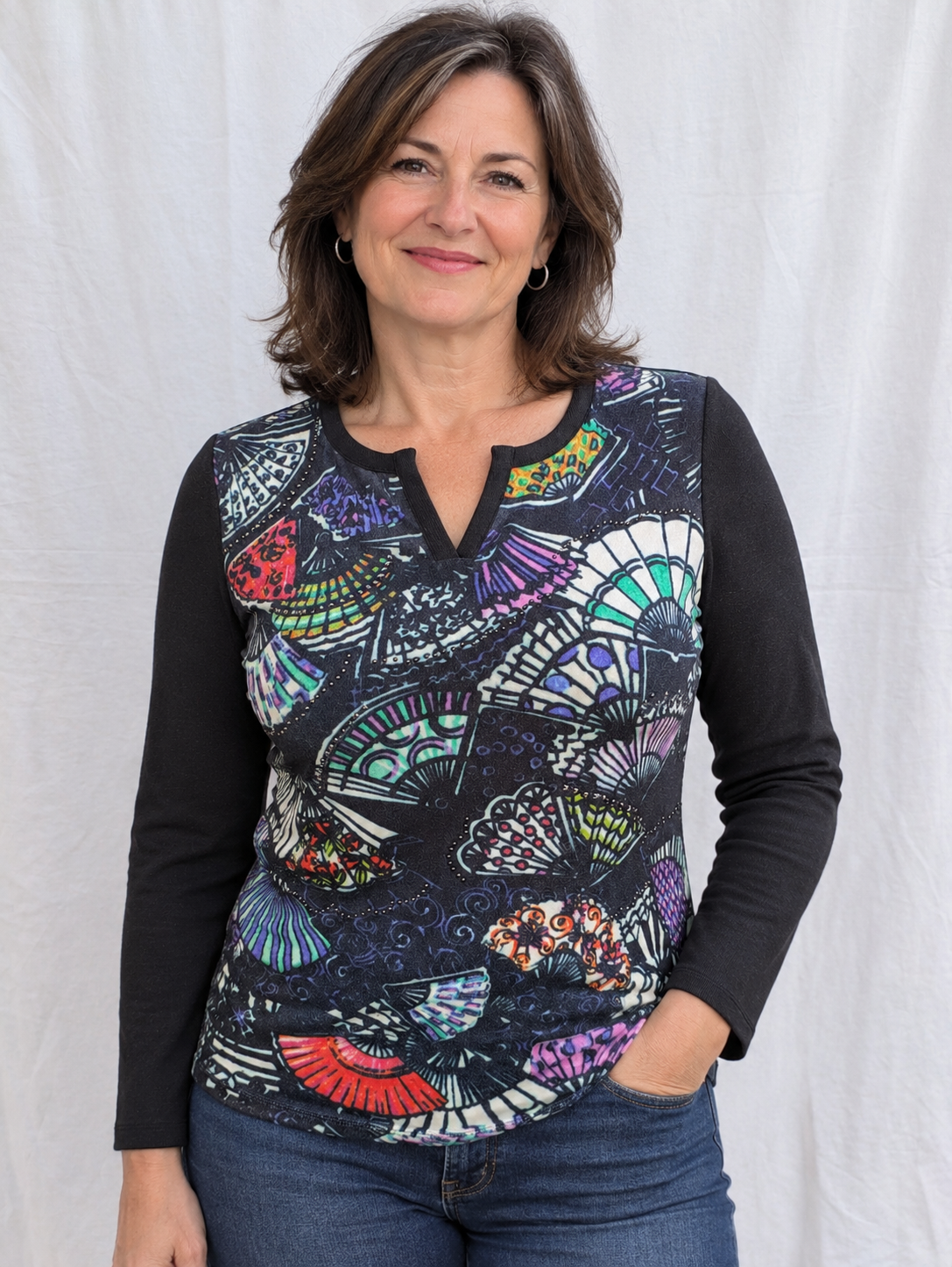 A woman modeling a long-sleeve henley top with a black base and a vibrant, multicolored abstract fan and wave pattern across the front, paired with blue jeans against a white fabric backdrop.